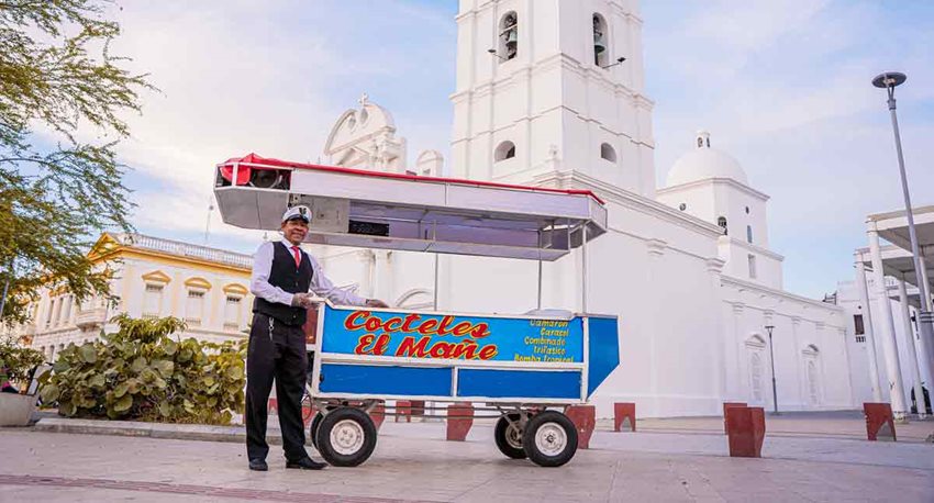 Hombre moreno de traje, posando al lado de su carro de comida de mar y detrás una iglesia blanca.