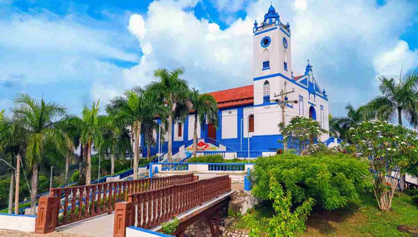 Foto de día de una iglesia blanco con azul, en medio de árboles y con un puente de madera enfrente.