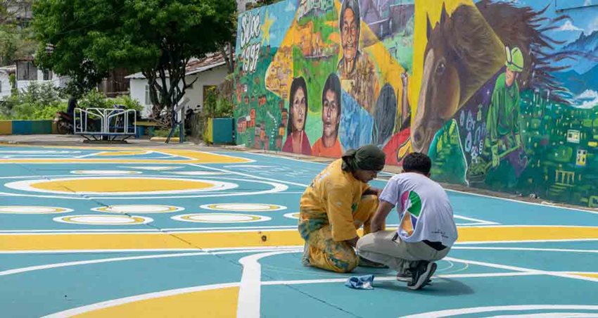 Dos hombres pintando el suelo de un parque, con un mural de personas y un caballo al fondo.