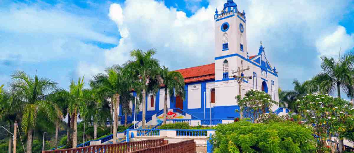 Descripción imagen: Foto de día de una iglesia blanco con azul, en medio de árboles y con un puente de madera enfrente.