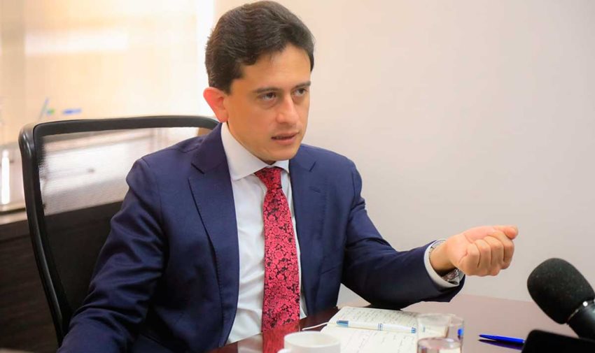 Ministro Luis Carlos Reyes, de traje y corbata roja, sentando en una silla hablando por micrófono.