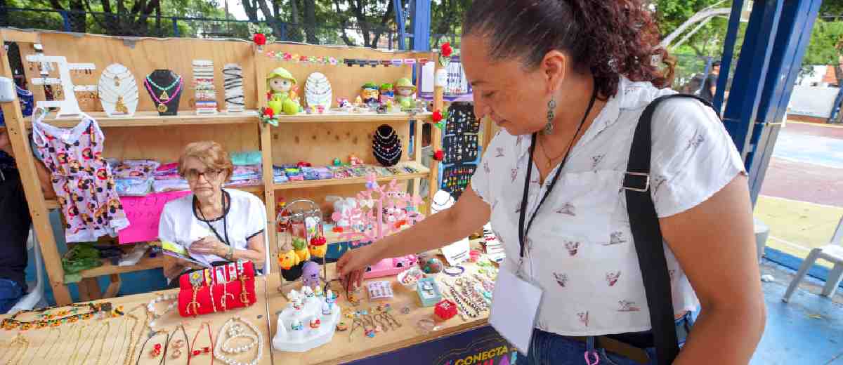 Descripción imagen: Mujer mirando artesanías en estand de la Feria Conecta Popular, mientras la señora del lugar la observa.