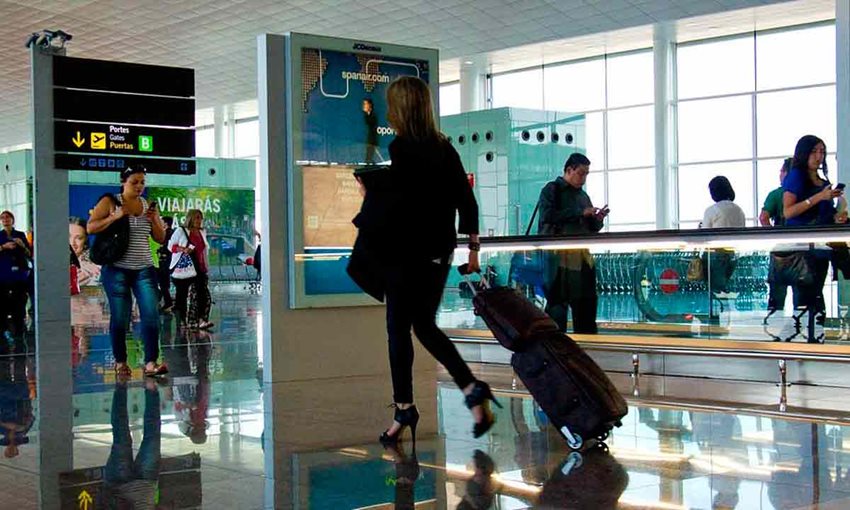 Mujer caminando con maleta de ruedas en medio de un aeropuerto, junto a personas alrededor que también caminan