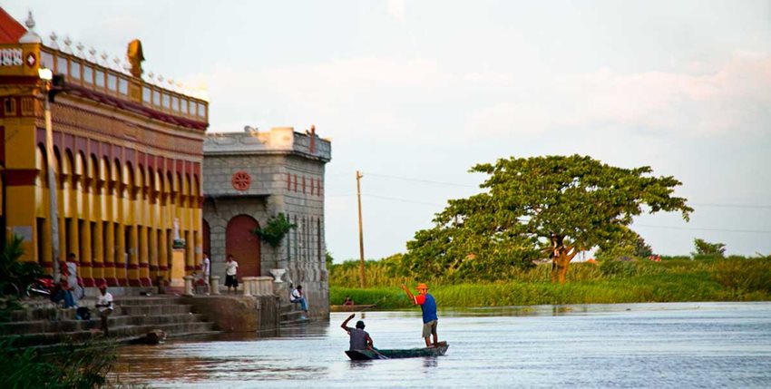 Al lado de dos edificios, dos hombres navegando en canoa por el río.