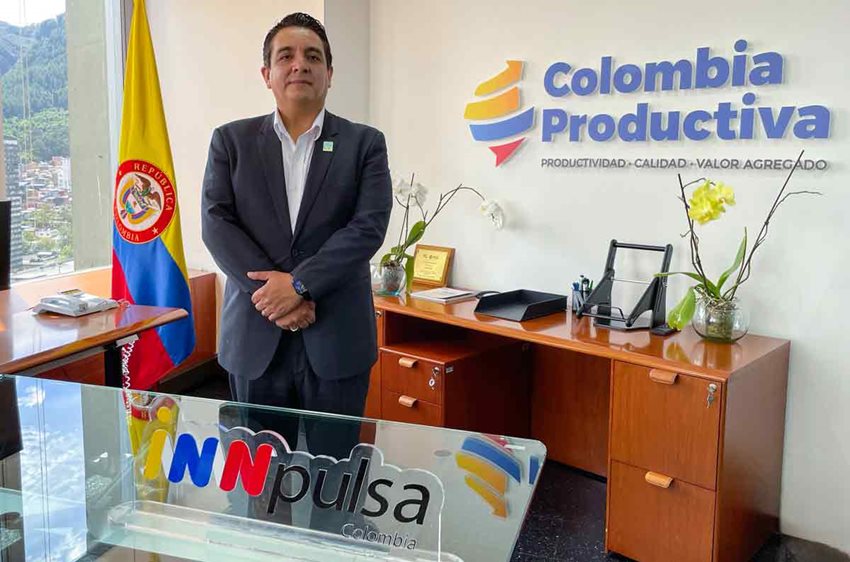 Hombre, de traje, posando para la foto de pie, con una bandera de Colombia detrás y logos de entidades al lado