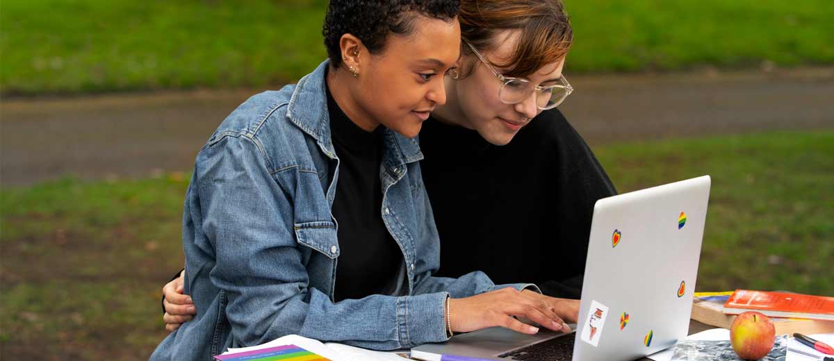 Descripción imagen: Dos mujeres abrazadas y sentadas, viendo un computador portátil blanco.