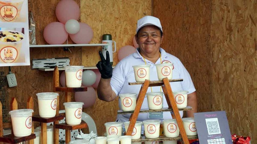 Mujer, vestida de blanco, posando sonriente para la foto al lado de su emprendimiento de postres.