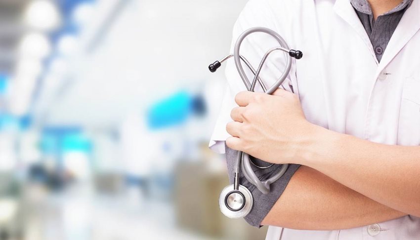 Médico, de frente, con bata blanca, los brazos cruzados y sosteniendo el estetoscopio en sus manos.