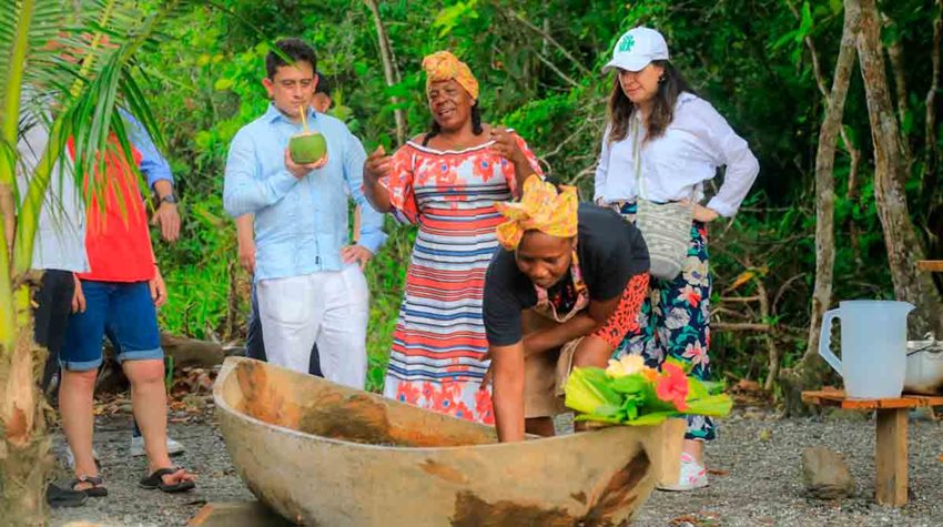 Hombre y mujeres hablando en medio de la vegetación y la arena, mientras observan balsa de madera.
