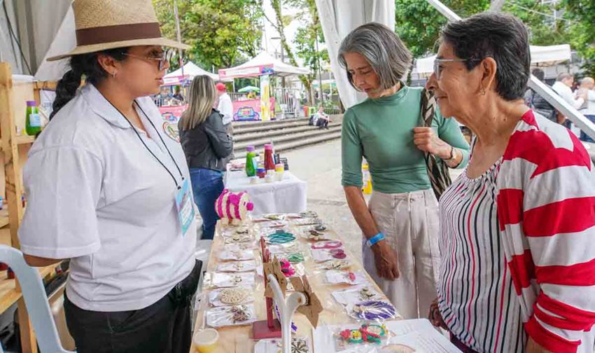 Mujeres de edad preguntando a mujer expositora sobre sus accesorios a la venta, en medio de la feria.