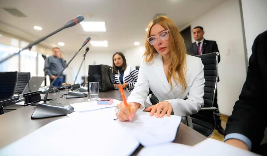 Ministra Diana Morales, con traje blanco y gafas, firmando documentos sobre una mesa.