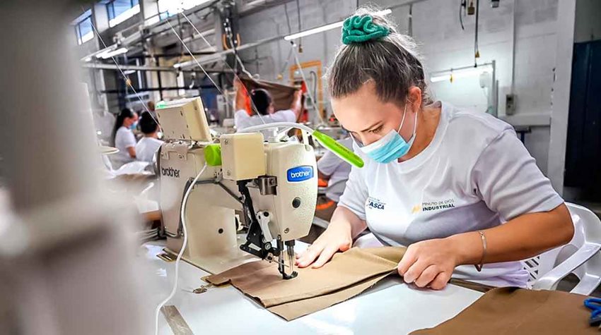 Mujer con el cabello recogido, camiseta blanca y tapabocas cosiendo en una máquina.