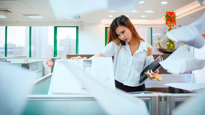 Mujer frente a una fotocopiadora, en una oficina, con papeles volando alrededor.