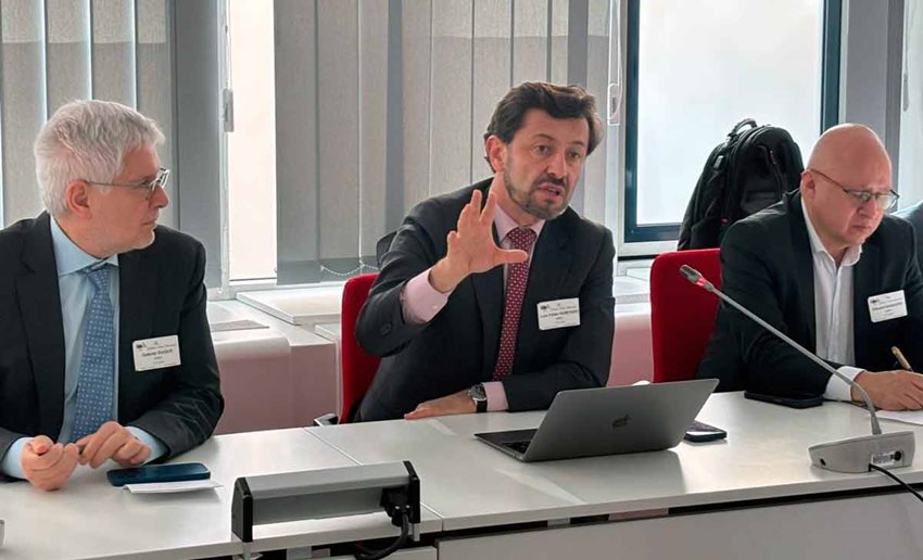 Tres hombres de traje, sentados frente a una mesa, y uno de ellos hablando y levantando una mano.