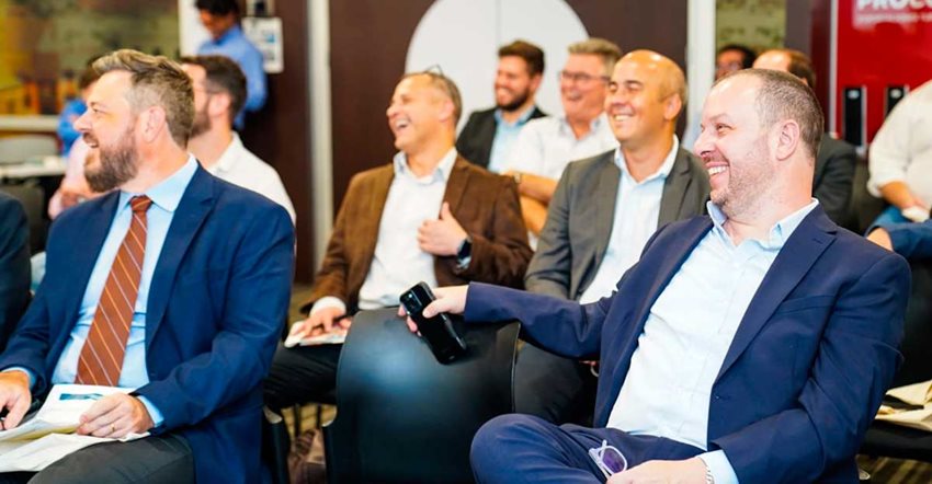 Hombres, de traje, sentados, viendo al frente y riéndose.