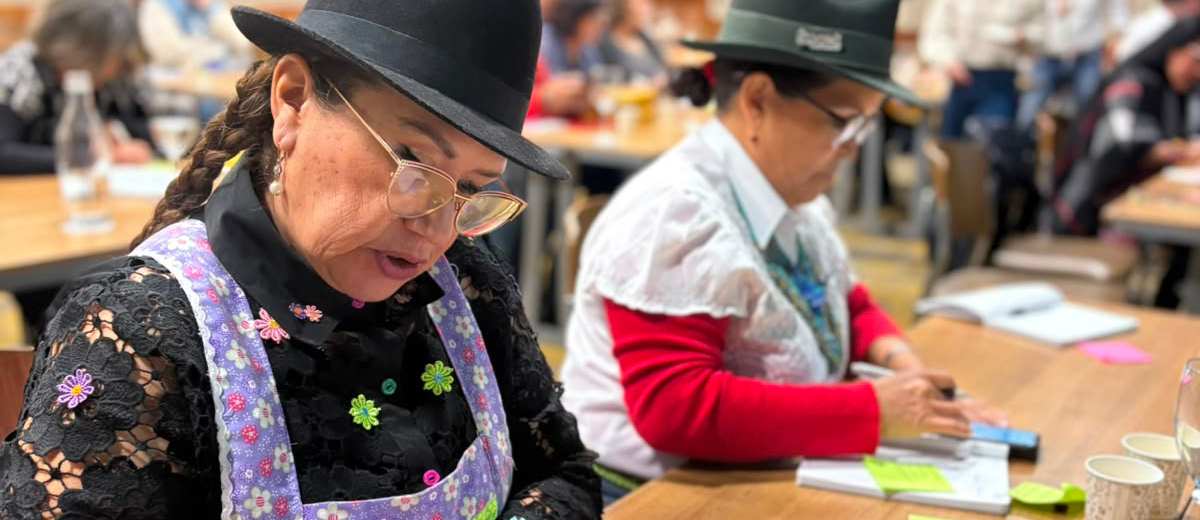 Descripción imagen: Mujeres vestidas de campesinas, escribiendo sobre papeles puestos en mesa.