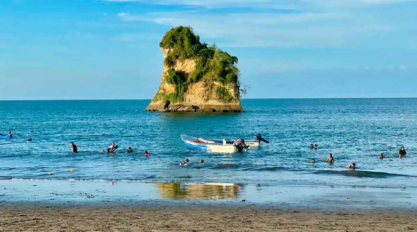 Fotografía de playa, mar con personas dentro y una roca grande al fondo.