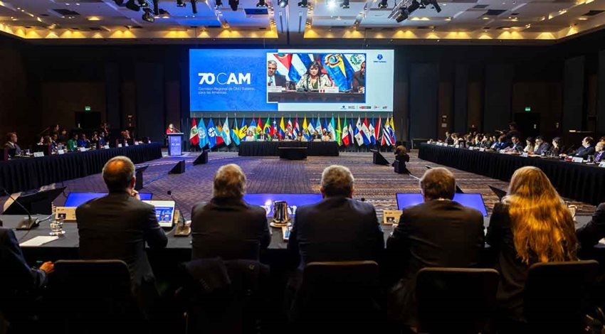 Foto de cuatro hombres y una mujer de espalda, sentados, viendo hacia el escenario y diapositivas al frente.