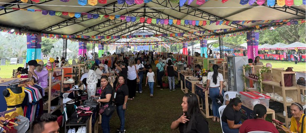 Descripción imagen: Personas caminando en medio de feria, con estands armados debajo de una gran carpa blanca, y viendo productos en venta.