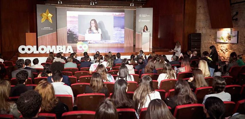 Personas sentadas en auditorio prestando atención a mujer hablando en el escenario.