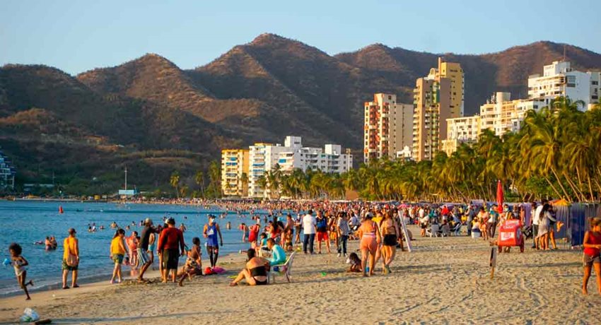 Personas caminando y sentadas en la playa, frente al mar, y en el fondo edificios y palmeras.