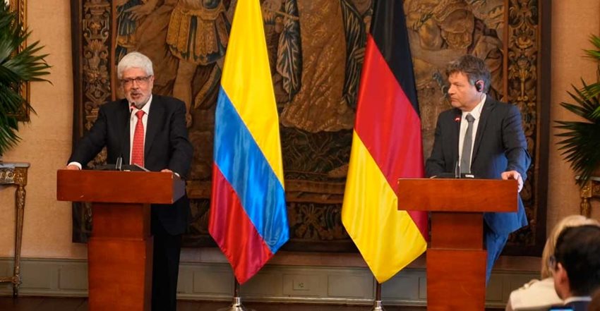 Dos hombres frente a dos atriles y en medio las banderas de Colombia y Alemania.