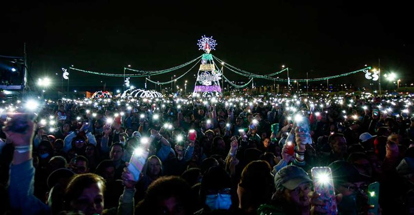 En el fondo, árbol grande de Navidad iluminado, mientras las personas lo rodean y alumbran con celulares.
