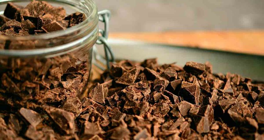 Fotografía de pedazos de chocolate sobre la mesa y dentro de recipiente transparente.