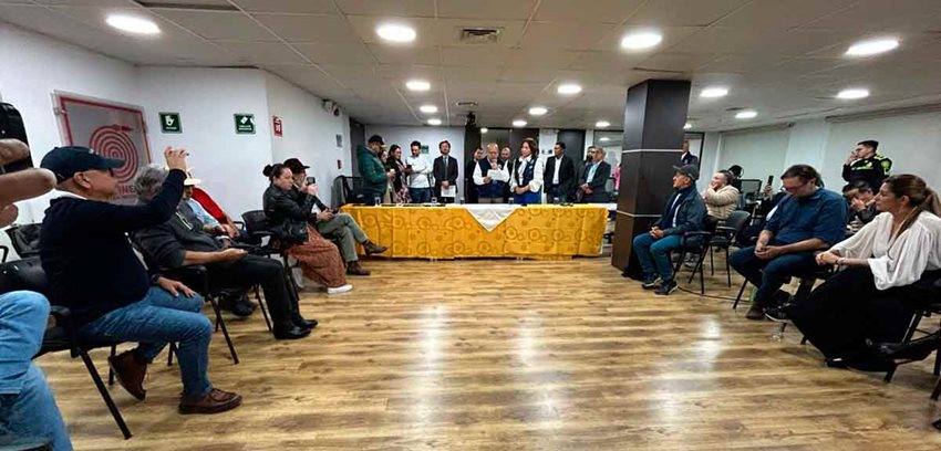 Personas reunidas en un salón, escuchando a hombre que está leyendo un documento por medio de un micrófono.