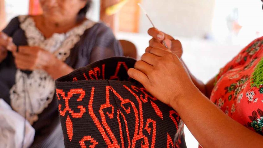 Manos de mujer tejiendo una mochila y al fondo otra mujer haciendo artesanías.