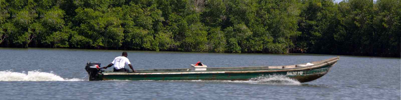 imagen de: Hombre moreno andando en lancha por medio del río y al fondo árboles.