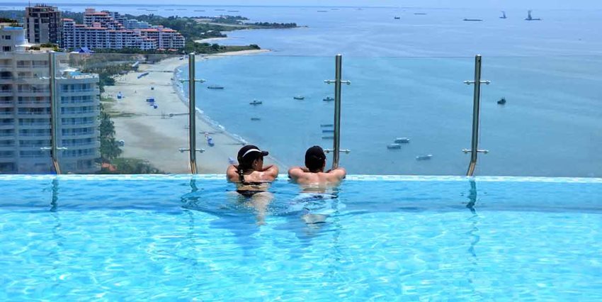 Mujer y hombre, con gorras, dentro de una piscina mirando hacia el mar y los edificios.