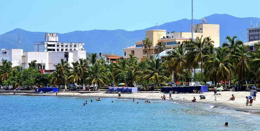 Foto panorámica, de día, de una playa de Santa Marta, con palmeras, personas y carpas azules alrededor.