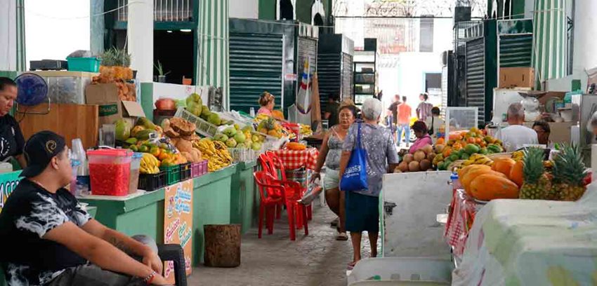 Personas caminando en medio de puestos de frutas en una plaza de mercado.