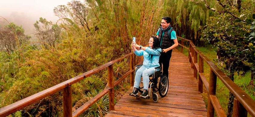 Hombre sosteniendo a mujer en silla de ruedas, mientras ella toma una foto al paisaje.