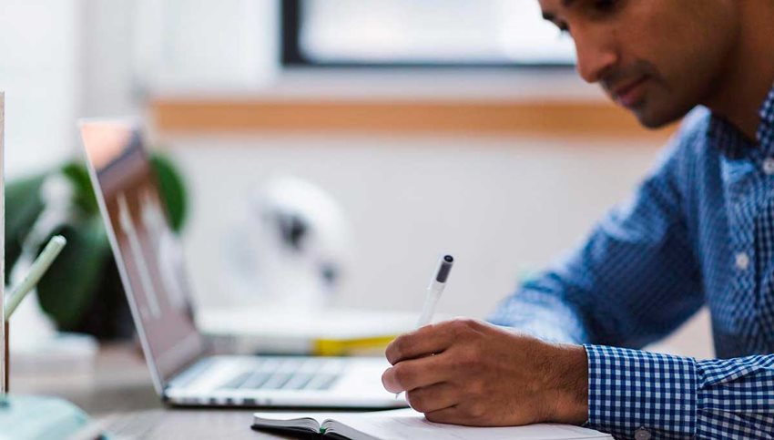 Hombre moreno escribiendo en cuaderno y con un computador portátil al frente.