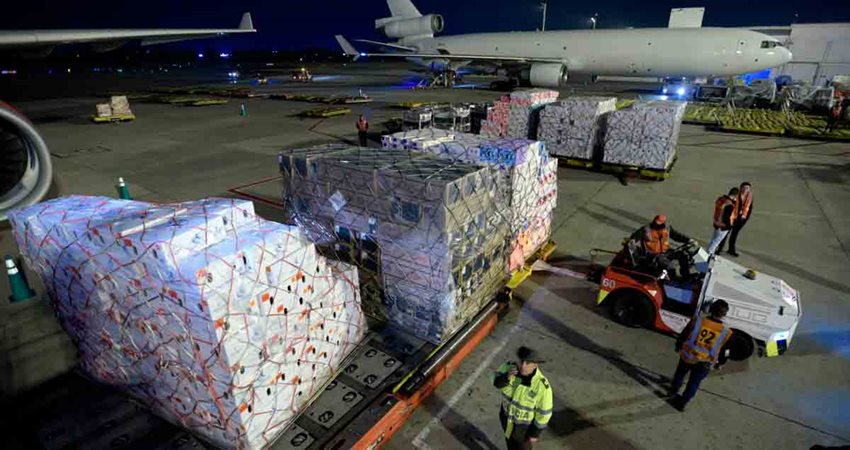 Mercancía descargada en medio de la pista de un aeropuerto, con un avión estacionado atrás.