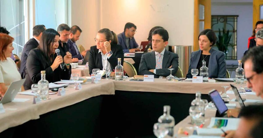 Mujeres y hombres sentados alrededor de una mesa y reunidos hablando por micrófono.