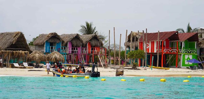 Foto del mar y al fondo cabañas, playa y una lancha estacionada, rodeada por boyas.