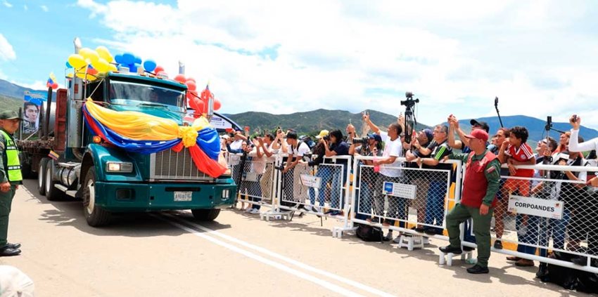 Camión verde con banderas de Colombia y Venezuela en el frente, pasando por un puente mientras la gente saluda