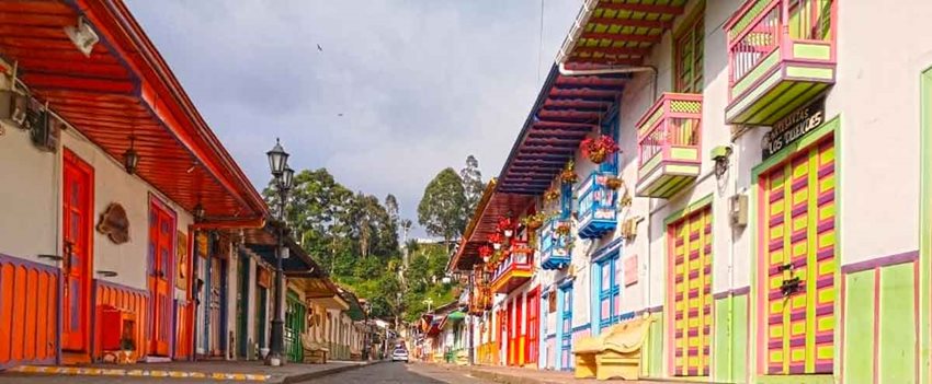 Foto de fachadas de casas de colores, a lo largo de una calle, y en el fondo unos árboles.