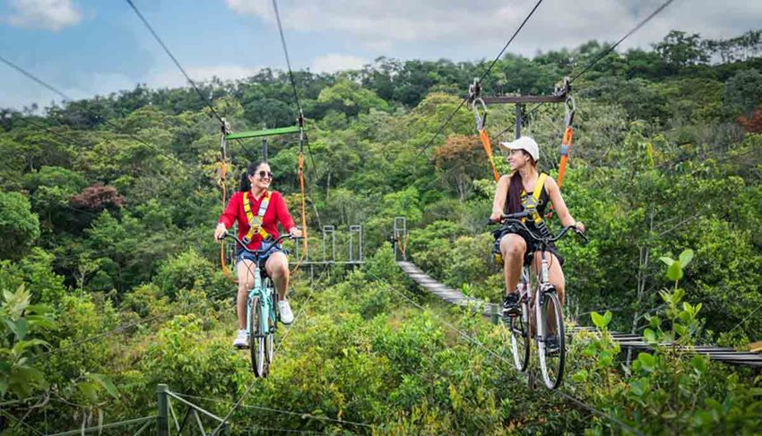 Dos mujeres montando bicicletas en el aire, sostenidas por cables, en medio de la naturaleza.