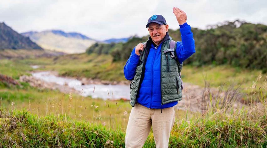 Hombre de la tercera edad, con ropa deportiva y gorra, saludando a la cámara en medio del campo.