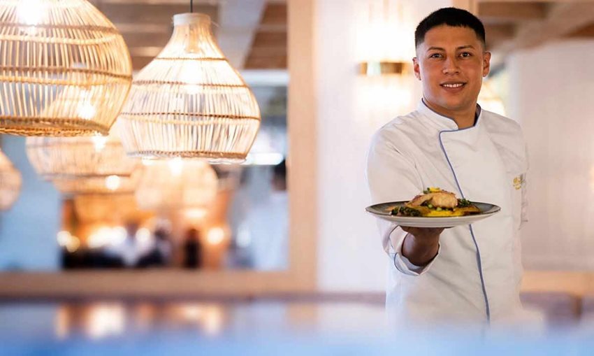 Descripción de la imagen: Hombre, con traje blanco de chef, sosteniendo en una mano un plato con comida y viendo a la cámara.