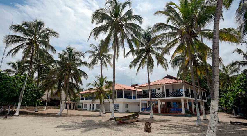 Descripción de la imagen: Casa blanca en medio de la playa y rodeada de palmeras y una lancha.