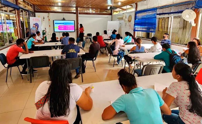 Personas acomodadas en mesas, prestando atención a una clase.
