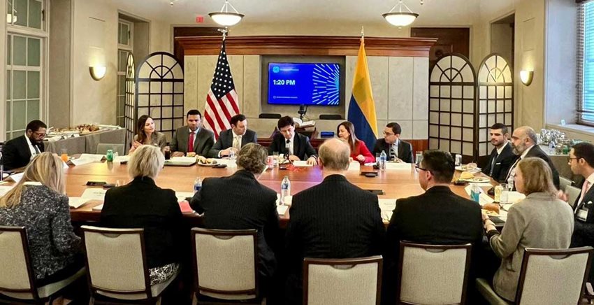 Personas sentadas alrededor de una mesa en una reunión, aparece una bandera de los Estados Unidos. 