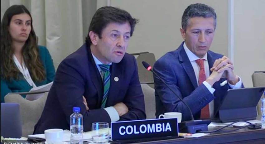 Dos hombres con traje, sentados, hablando por un micrófono a otras personas.