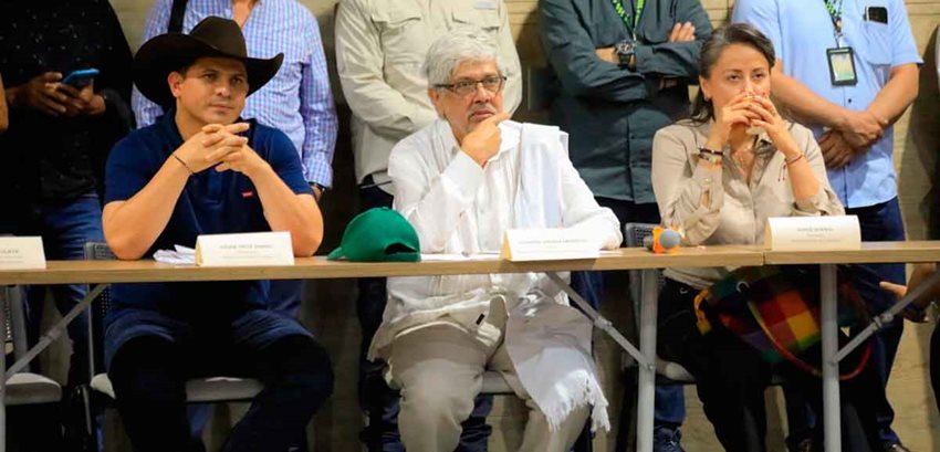 Dos hombres y una mujer en una mesa, sentados en sillas, prestando atención al público.