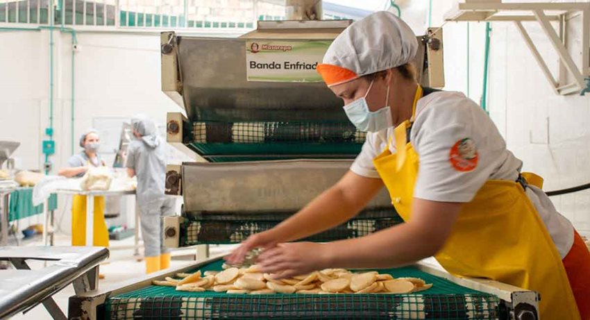 Mujer con uniforme y tapabocas trabajando en máquina procesadora de alimentos.